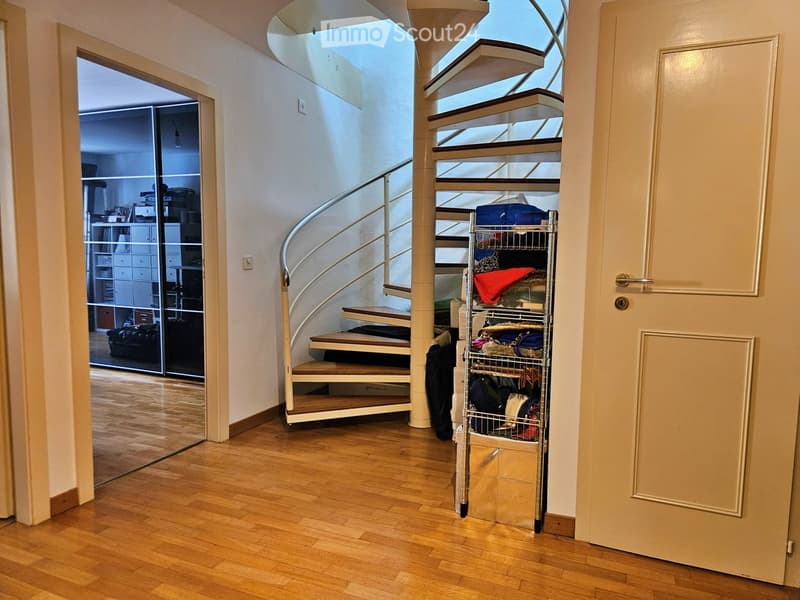 Spacious hallway with wooden flooring, curved staircase with glass railing, open door leading to another room, and a metal storage cart with various items.