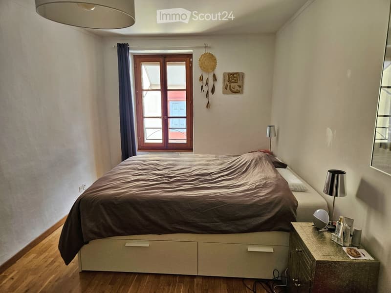Cozy bedroom with a large bed covered in dark brown bedding, a nightstand with a lamp, a mirror, and a window with curtains. The room features wooden flooring and a wall decoration.