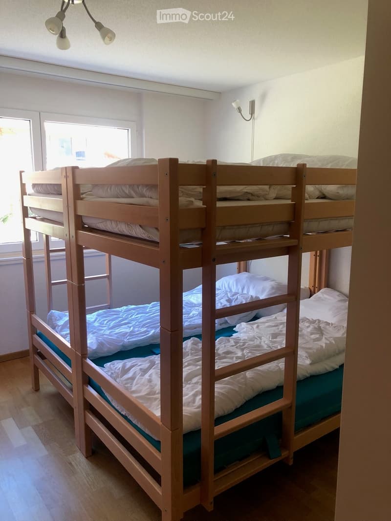 A cozy bedroom with two wooden bunk beds, each equipped with pillows and blankets. The room features light-colored walls, wooden flooring, and a window allowing natural light to enter.