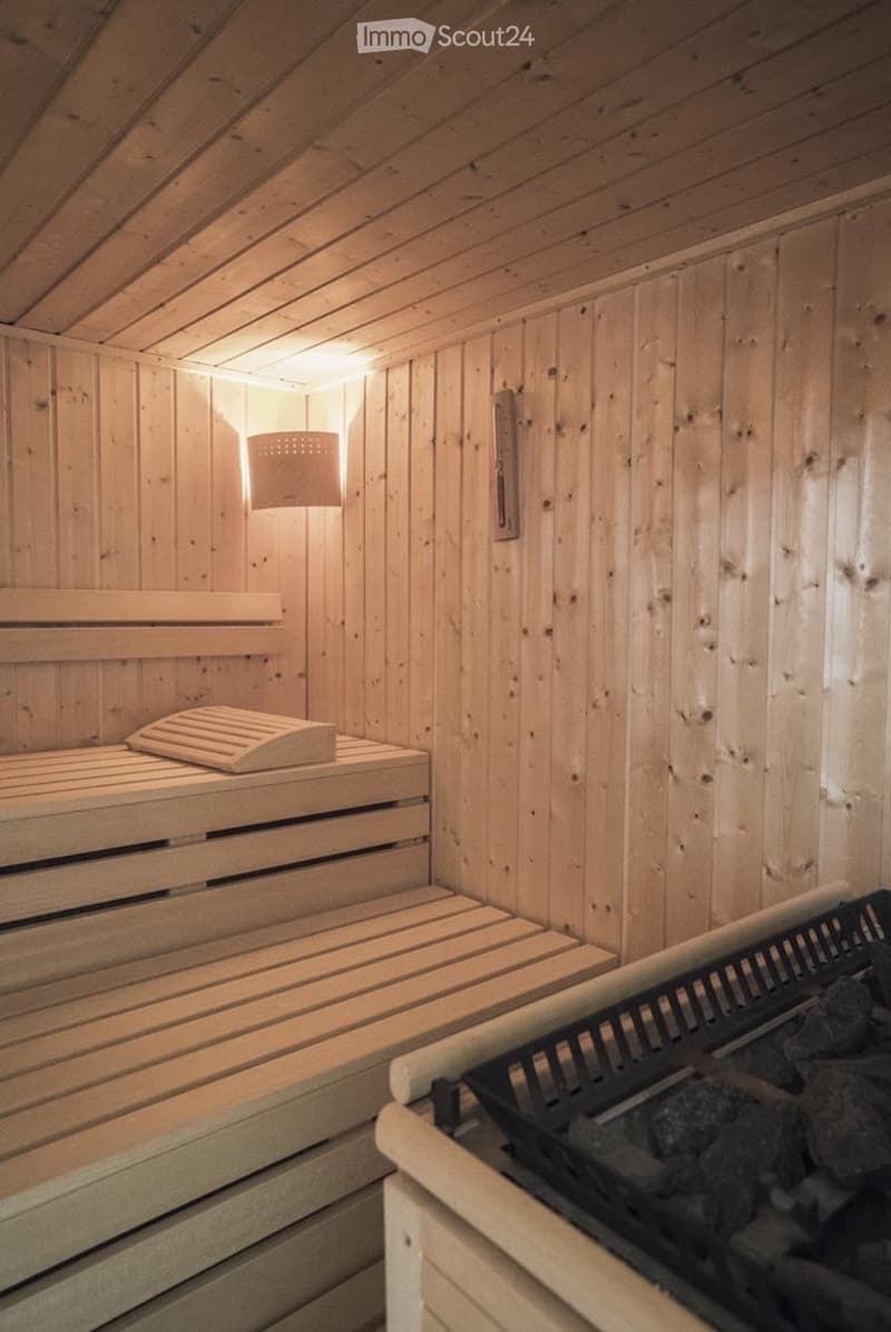A wooden sauna room with benches, a seat, a lamp, and a stone stove. The walls and ceiling are made of wood, creating a warm and cozy atmosphere.