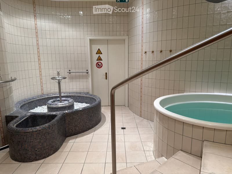 Spa area with a tiled shower stall and a bathtub, featuring a tiled floor and walls, a metal handrail, and a door with safety signs.