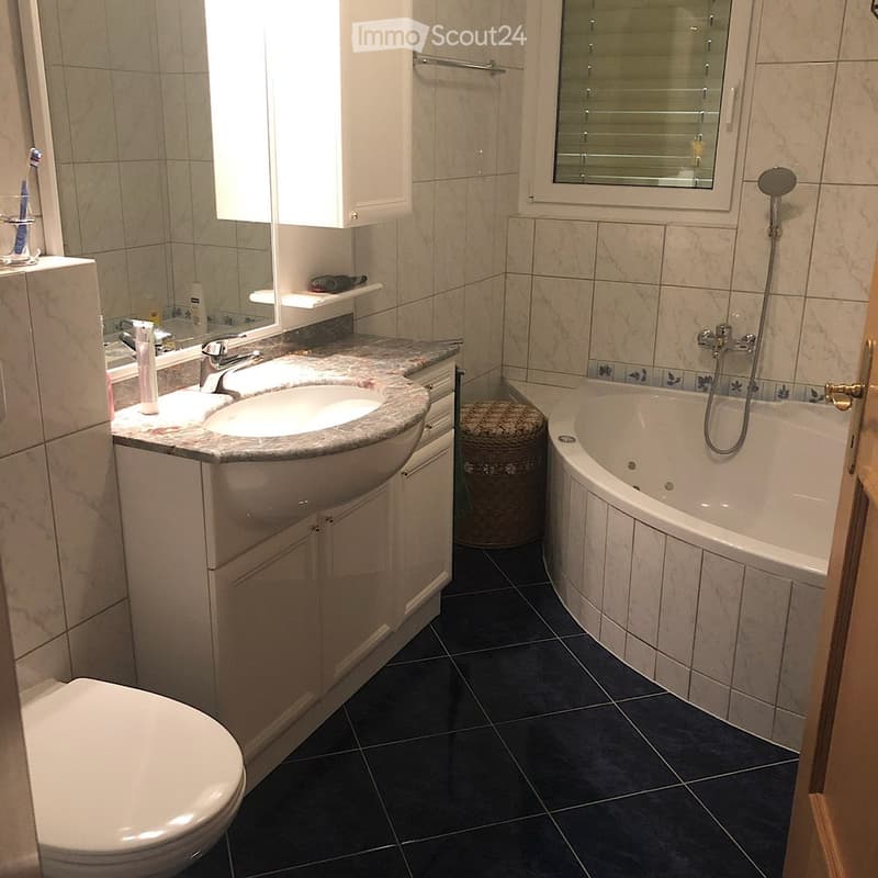 Modern bathroom with a double sink, toilet, bathtub, and shower head. Features include a marble countertop, dark tiled floor, and a window with blinds.