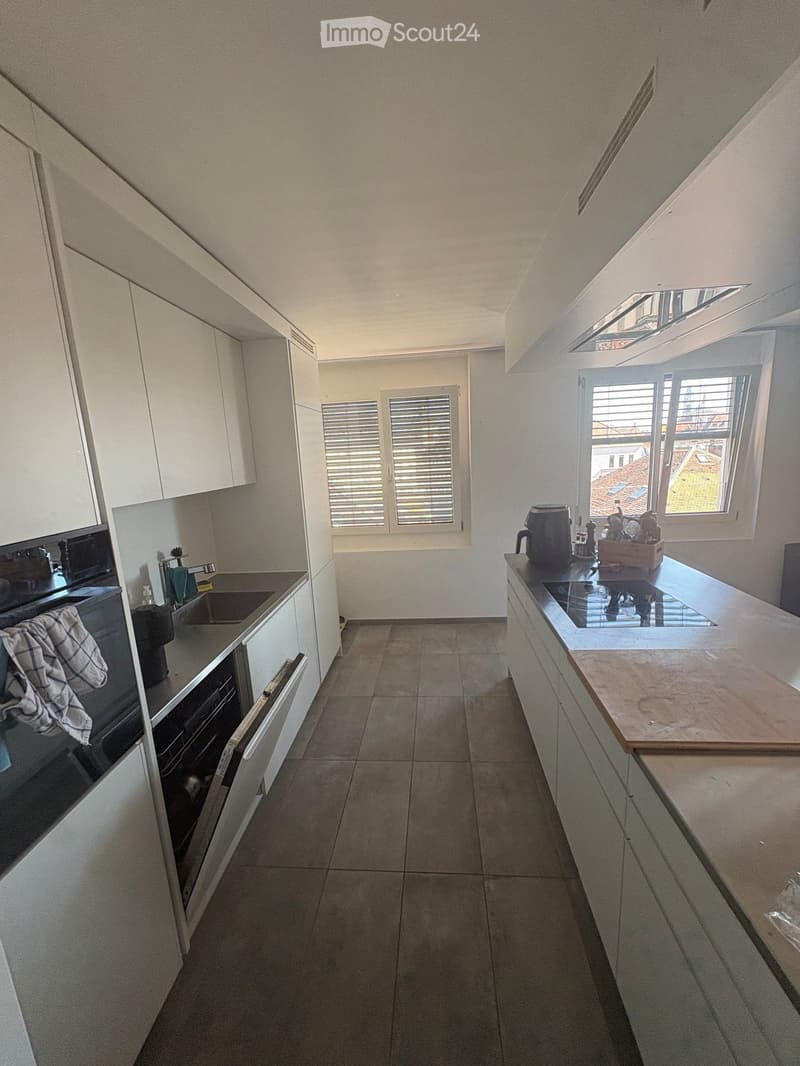 Modern kitchen with white cabinets, stainless steel appliances, and a central island with a wooden cutting board. The space features a tiled floor, large windows with blinds, and a sleek design.