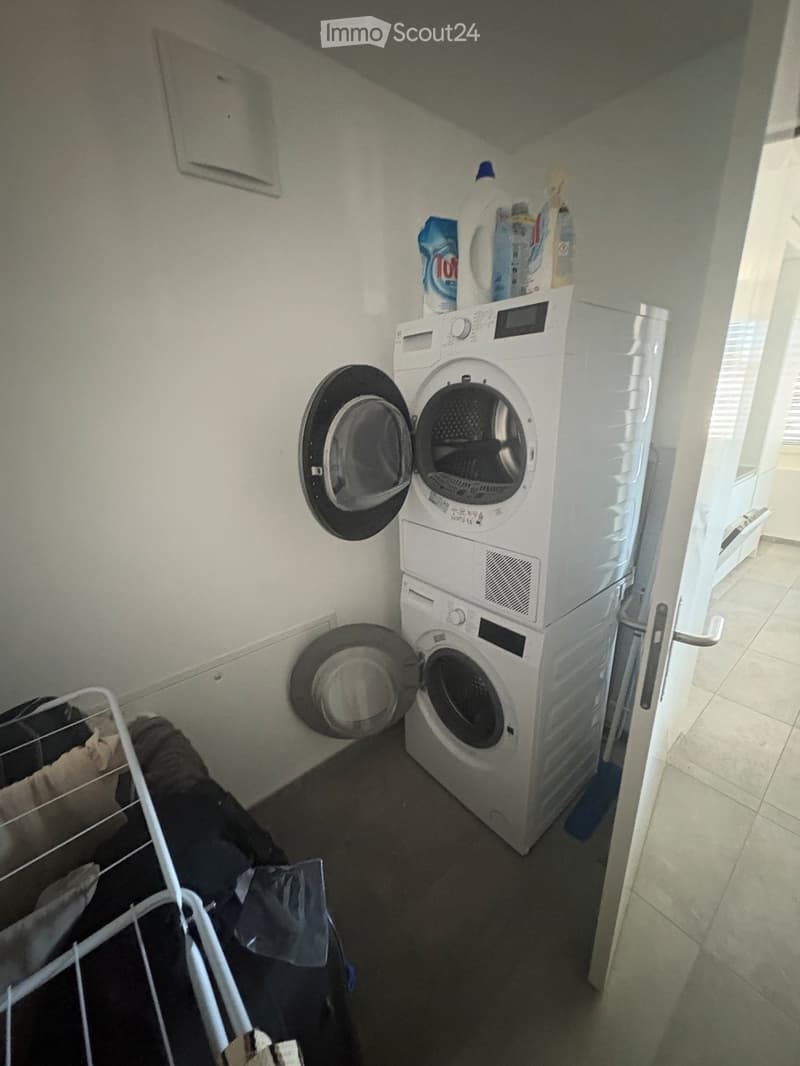 A small laundry area with a stacked washing machine and dryer, a hat hanging on the wall, and a laundry basket with clothes.