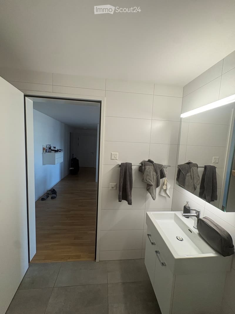 Modern bathroom with white tiles, a sink with a modern faucet, a large mirror with built-in lighting, a towel rack with several towels, and a small cabinet. The floor is tiled, and the walls are white.
