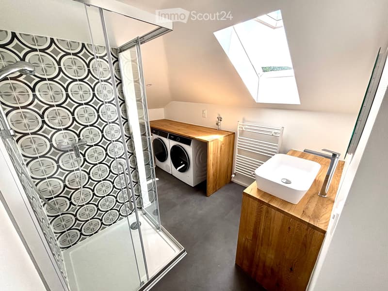Modern bathroom with a glass-enclosed shower, wooden vanity with sink, washing machine, radiator, and skylight window.