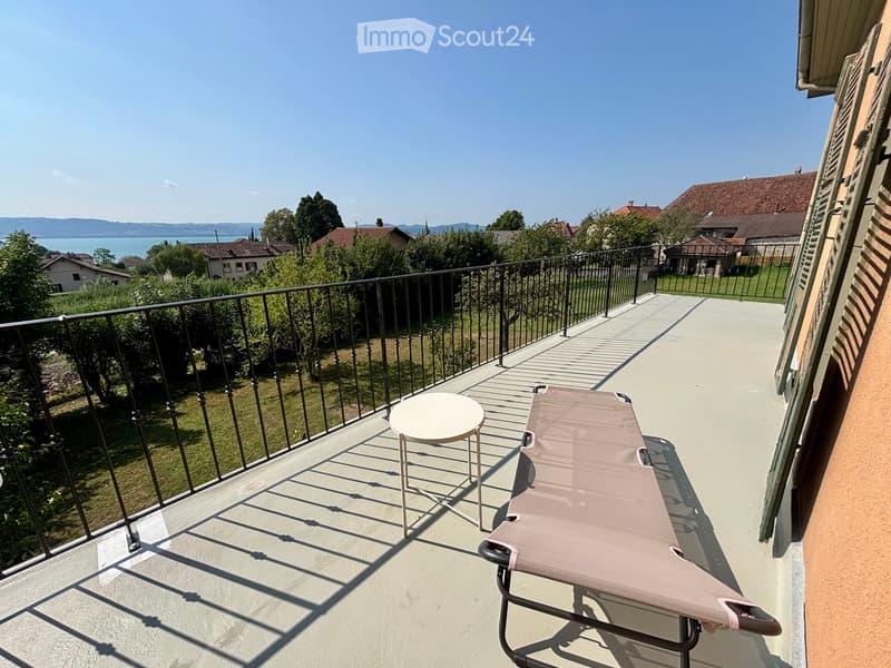 Balcony with a metal railing, a small round white stool, and a pink sun lounger, overlooking a scenic view of a lake and surrounding houses.
