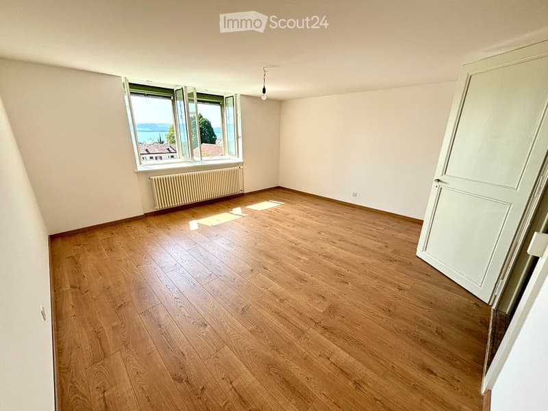 Empty room with wooden flooring, large window with view, radiator, and white walls.