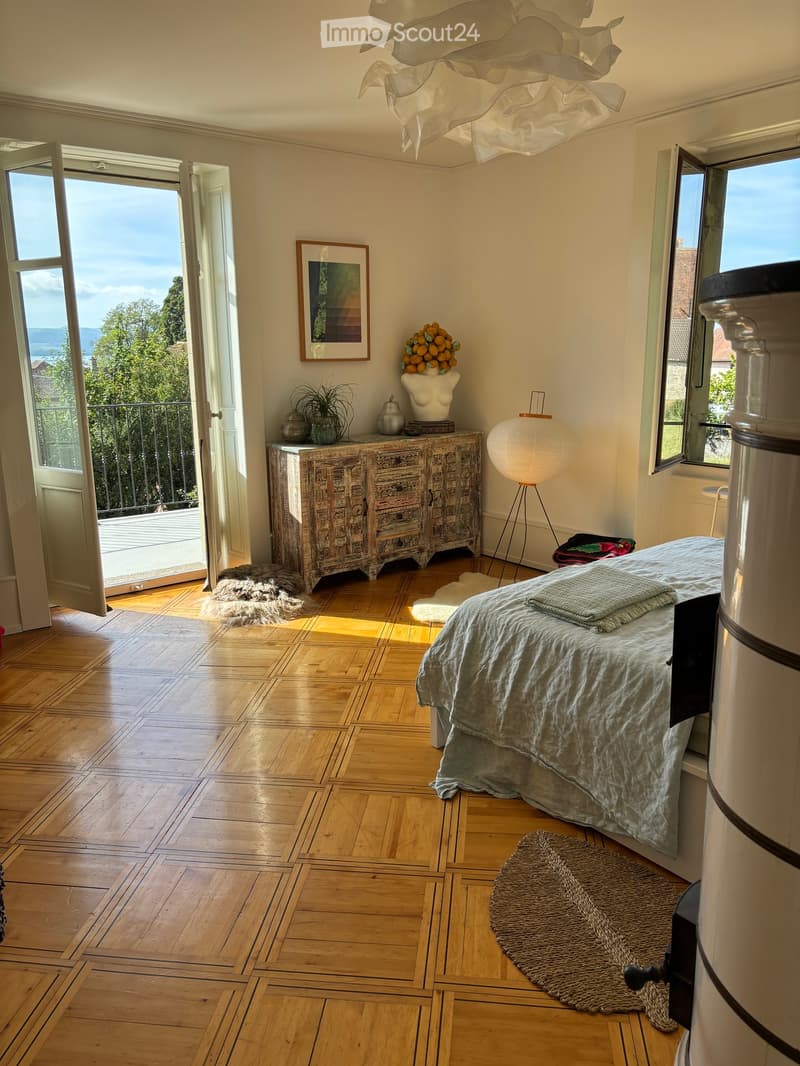 Bright bedroom with wooden floor, large windows, bed with grey bedding, cabinet, floor lamp, decorative items, and view of greenery.