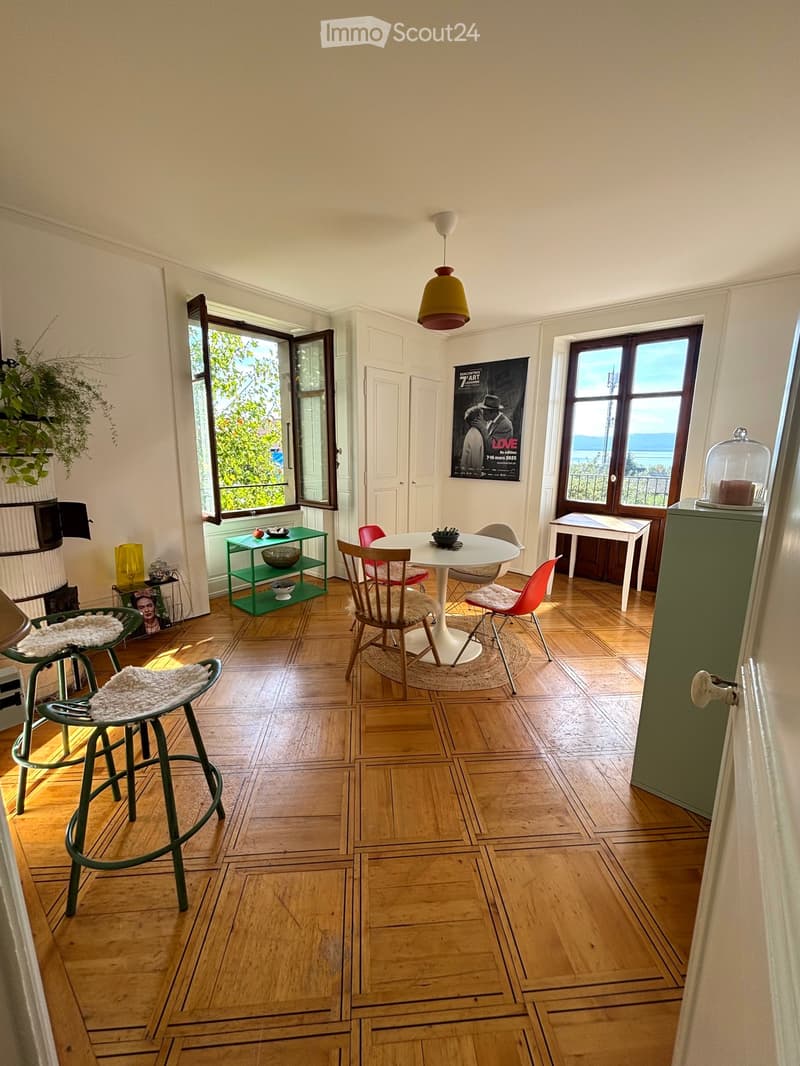 Brightly lit room with wooden flooring, large windows, a round white dining table with four chairs, a green shelf, and a hanging lamp.