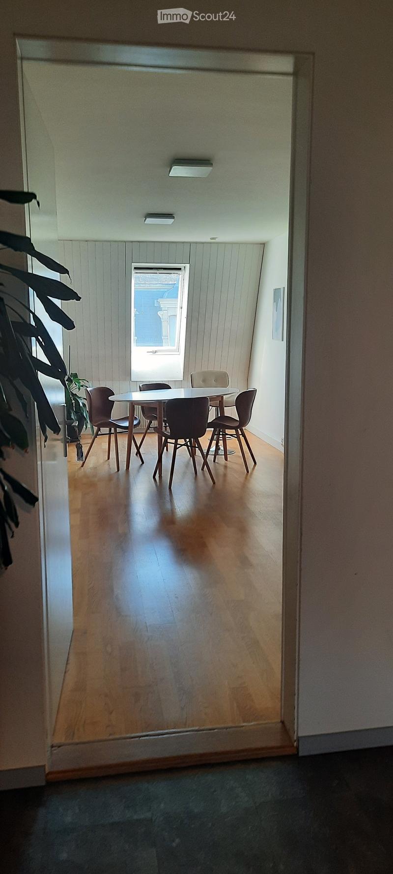 A small, well-lit room with wooden flooring, a round table surrounded by six chairs, and a window letting in natural light.