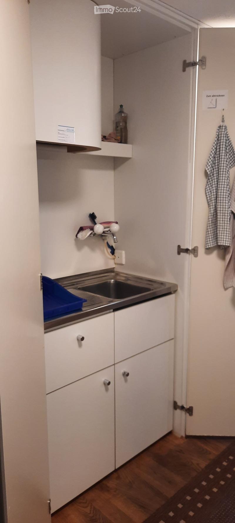 A compact kitchen with white cabinets, a stainless steel sink, and a wooden floor. A bottle is placed on the counter, and a towel hangs on the wall.