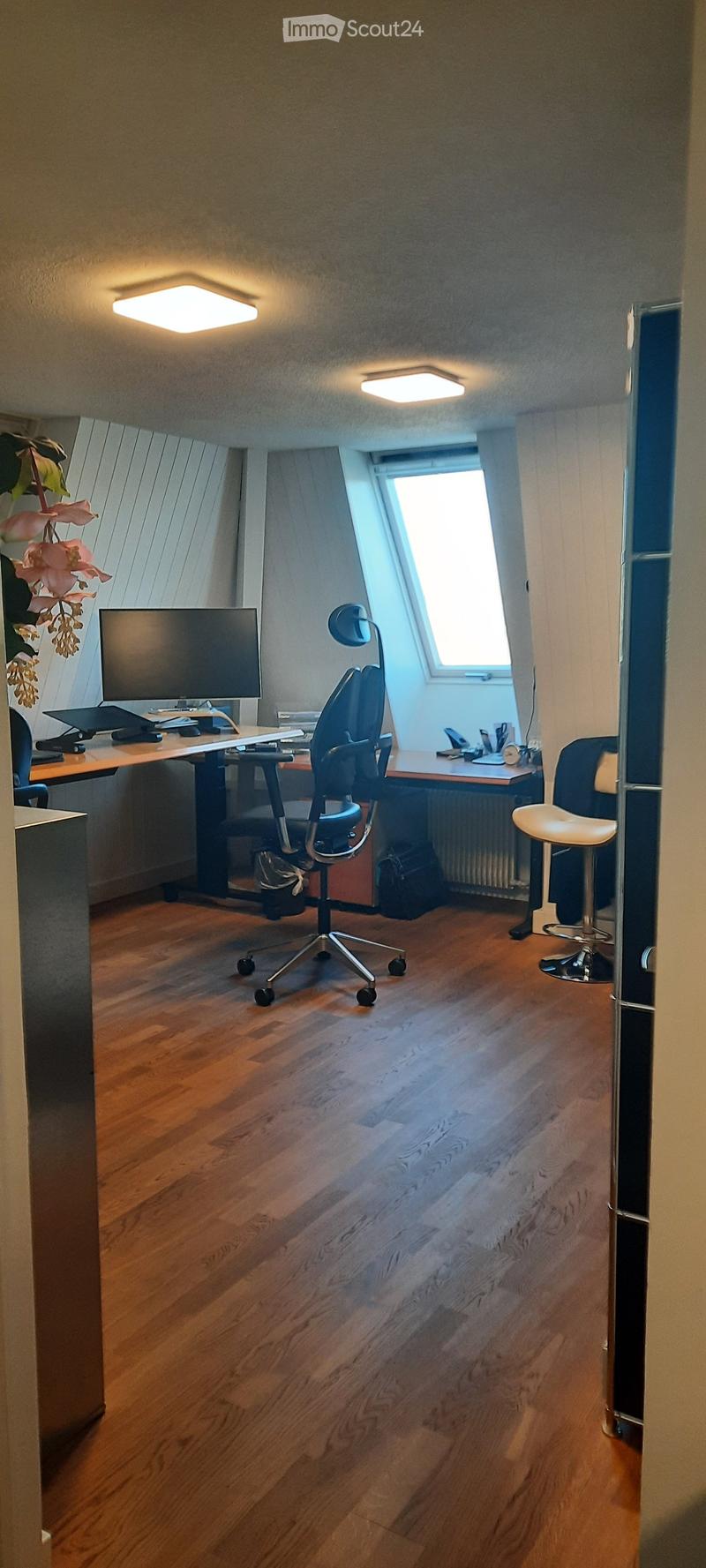 A well-lit office space with wooden flooring, a large window, a desk with a screen and laptop, a comfortable chair, a stool, and a decorative flower arrangement.