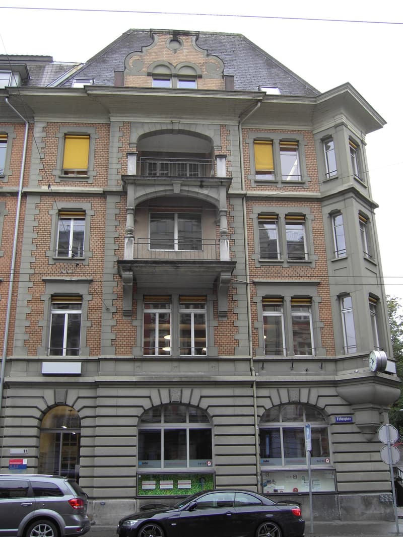 Four-story brick building with arched windows, balconies, and decorative stonework. Features a central entrance with large glass doors and a small green area in front.