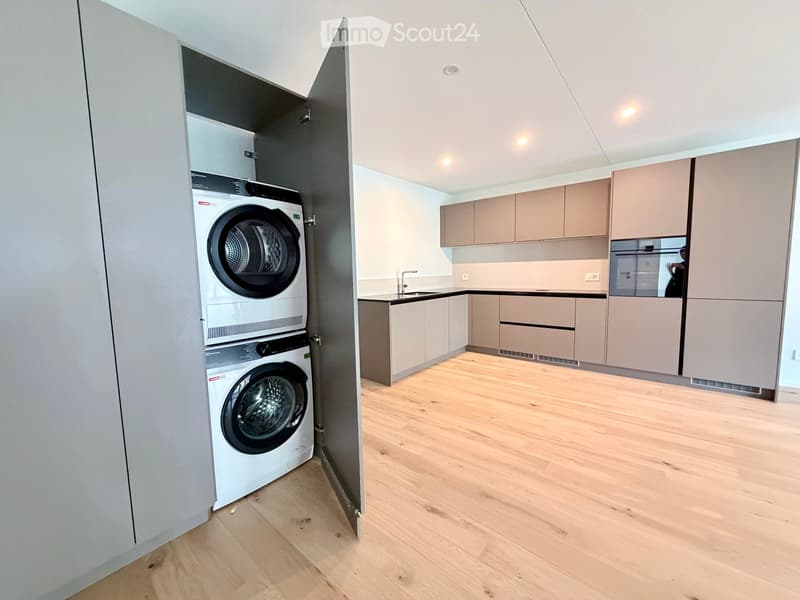 Modern kitchen with light wood flooring, light-colored cabinets, and a built-in dishwasher. A private washing machine is visible in an adjacent space.