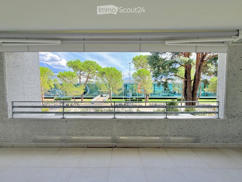 Balcony with a metal railing, tiled floor, and a view of a landscaped area with trees and a modern building in the background.