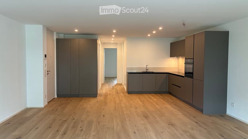 Empty kitchen with wooden flooring, grey cabinets, and built-in appliances, including a sink and oven. The space is well-lit with recessed lighting.