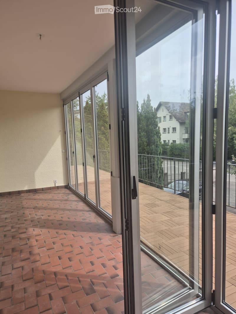 Spacious balcony with brick flooring, large glass sliding doors, and a view of a residential area with trees and a house.