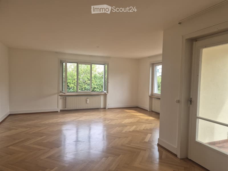 Empty room with wooden flooring, white walls, and large windows allowing natural light. Radiators are visible under the windows.