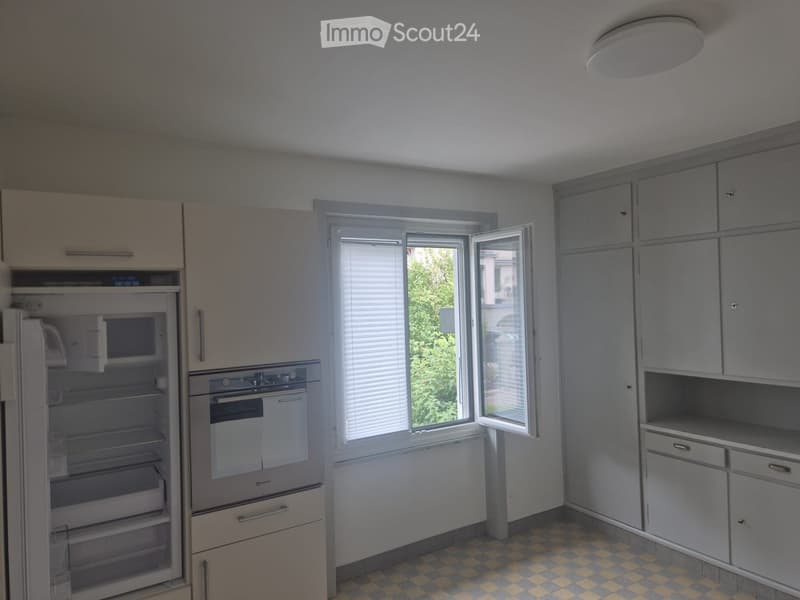Modern kitchen with grey cabinets, a refrigerator, microwave, and an open window with blinds.