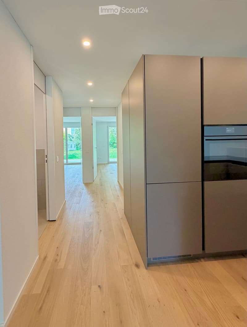 Bright, modern hallway with light wooden flooring, white walls, and recessed lighting. Features built-in cabinets and a stainless steel appliance on the right side, with open doors leading to other rooms.