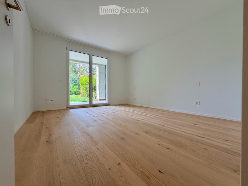 Empty room with light wooden flooring, white walls, and large sliding glass doors leading to a garden.