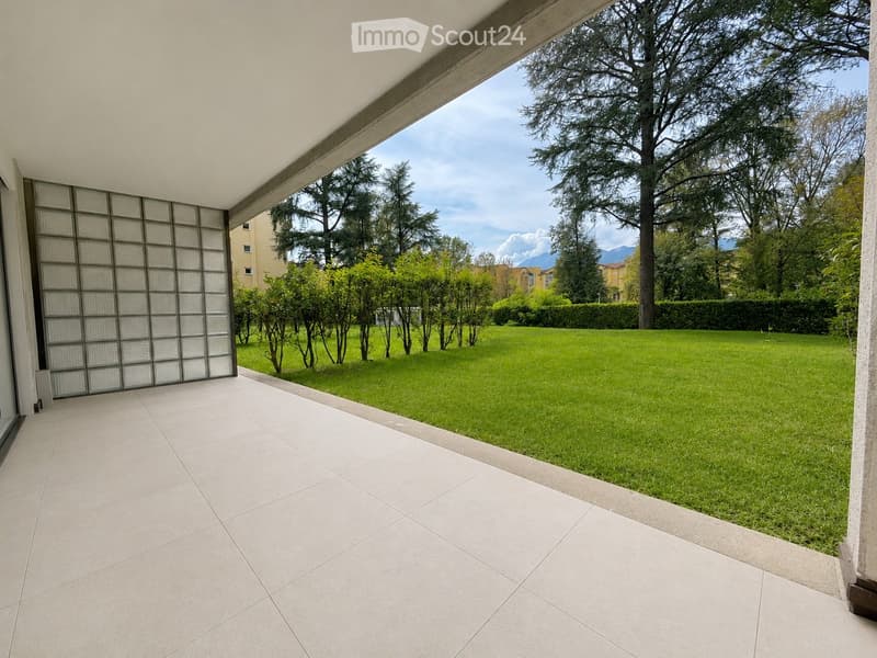 A spacious outdoor area with a tiled patio leading to a well-maintained lawn, surrounded by trees and greenery, with a view of distant mountains.