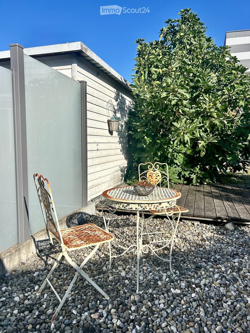 A small outdoor seating area with a round metal table and two matching chairs on a gravel surface, adjacent to a wooden deck and a modern building with a glass wall.
