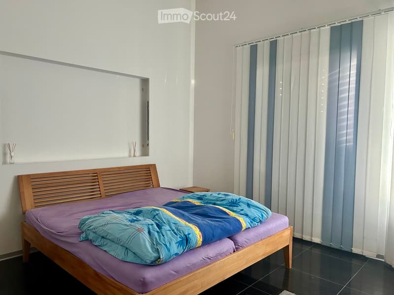 A modern bedroom with a wooden bed frame, purple bedding, and a blue floral duvet. The room features white walls, dark tiled flooring, and a window with white and blue curtains.