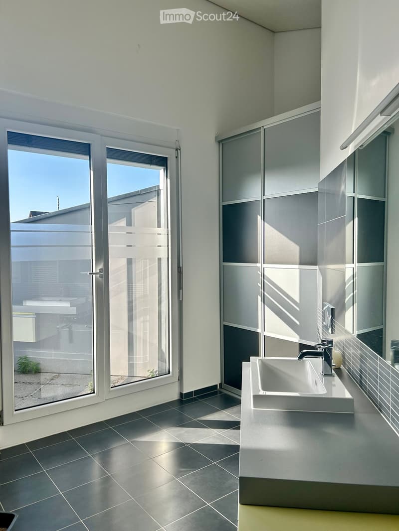 Modern bathroom with dark tiled floor, white sink, sleek faucet, large windows, and a view of the outdoor area.