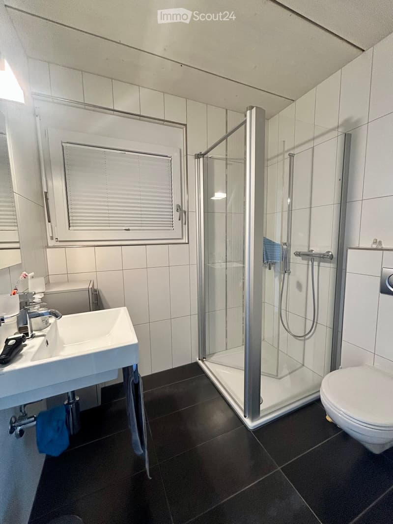 Modern bathroom with white tiles, a glass-enclosed shower, a white sink, and a toilet. Blue towels are placed on the floor and near the sink.