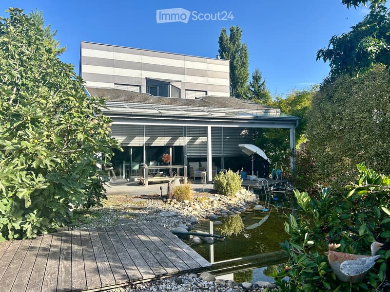 Modern house with a spacious garden featuring a small pond, wooden decking, and surrounding greenery.