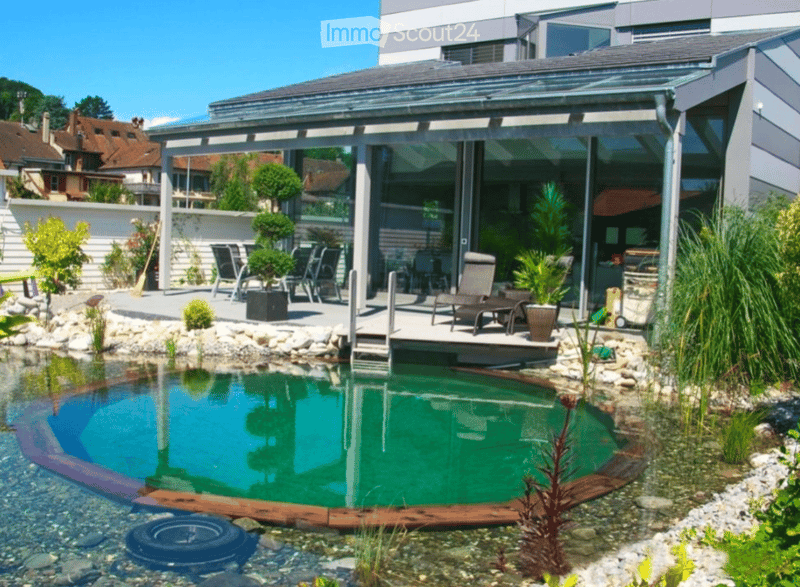 Modern outdoor area with a small swimming pool, surrounded by rocks and plants, featuring a covered patio with lounge chairs and a view of the surrounding neighborhood.