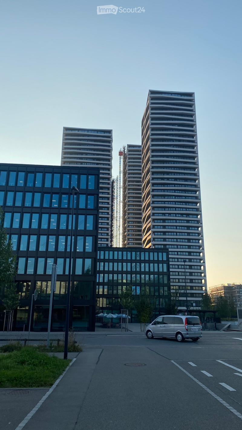 Modern high-rise residential buildings with glass facades and balconies, surrounded by a paved area with a parked van and some outdoor seating.