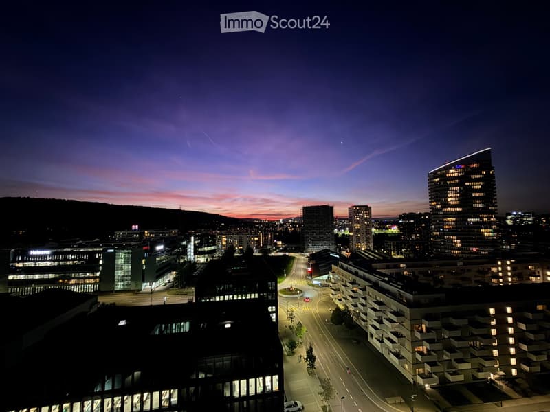 A cityscape at dusk with modern high-rise buildings, illuminated windows, and a winding road.