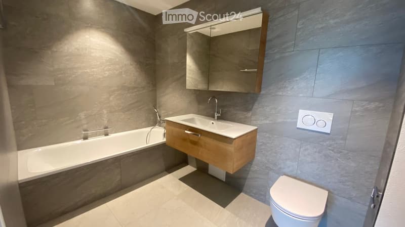 Modern bathroom with a bathtub, sink, mirror, and toilet. Features grey tiled walls and a wooden vanity.