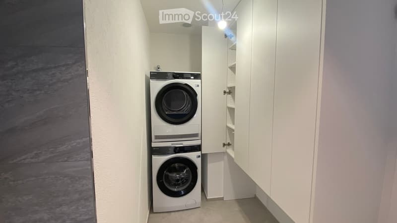 A small, well-lit room with white walls and a light-colored floor, featuring a stacked washer and dryer unit with a built-in cabinet for storage.
