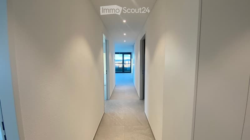 A modern, well-lit hallway with white walls, tiled flooring, and recessed ceiling lights. The corridor features a window at the end, allowing natural light to enter.
