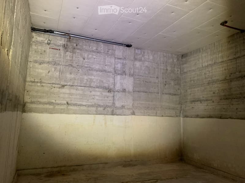 Empty room with concrete walls and a white drop ceiling, showing exposed pipes and a light fixture.