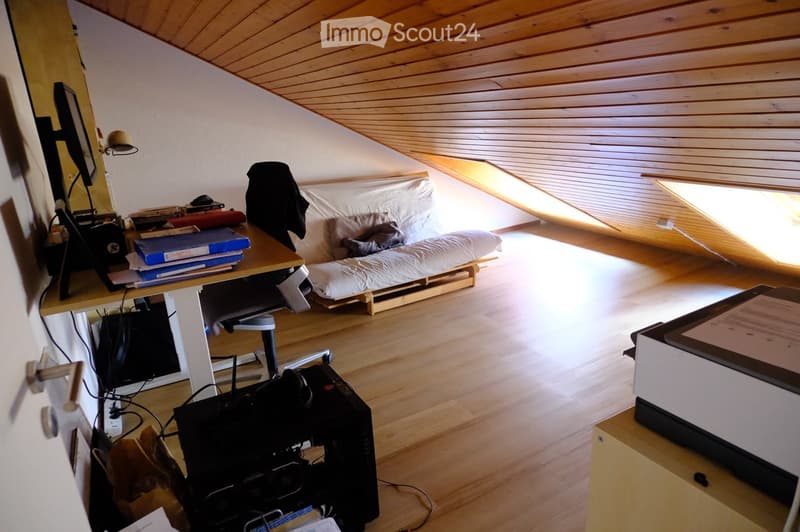 A cozy attic room with wooden floors and slanted wooden ceilings. Features include a desk with a laptop, a small sofa, a printer, and a lamp. The room has large skylights allowing natural light to fill the space.