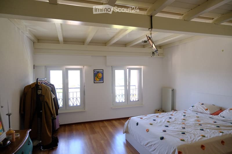 Bedroom with wooden floor, white walls, exposed wooden beams, large windows with metal grilles, bed with colorful stones on the blanket, nightstand, radiator, and coat rack with hanging coats.