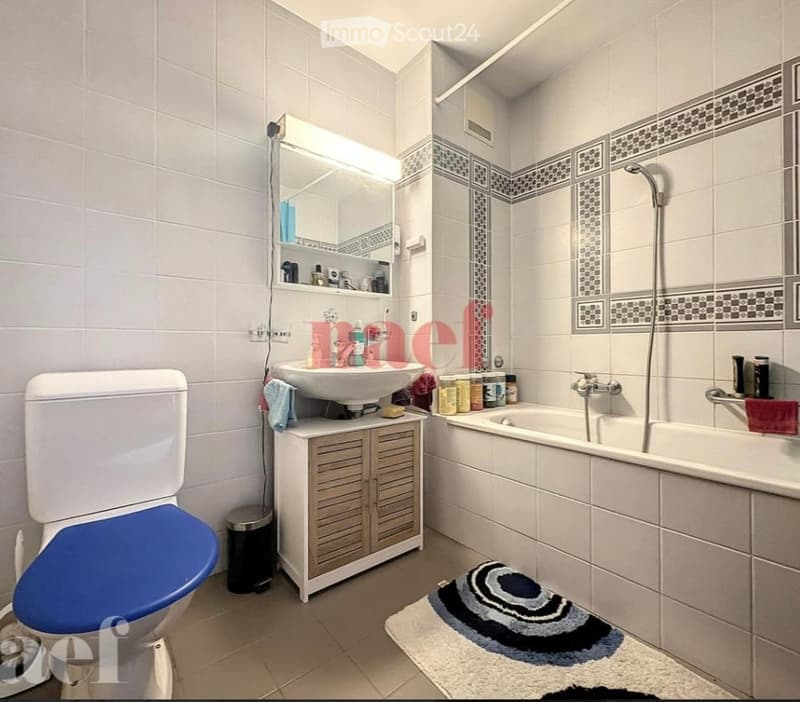 Modern bathroom with white tiles, a toilet with a blue seat cover, a sink with a cabinet below, a bathtub with a showerhead, a mirror with shelves, and a bathmat on the floor.