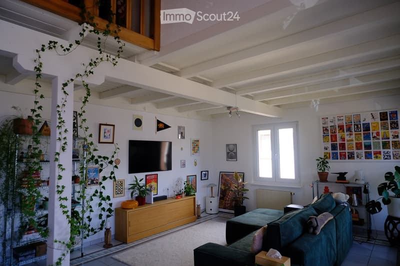 Modern living room with a green sofa, coffee table, television, and several potted plants. The room features white walls, exposed wooden beams, and large windows allowing natural light to fill the space.