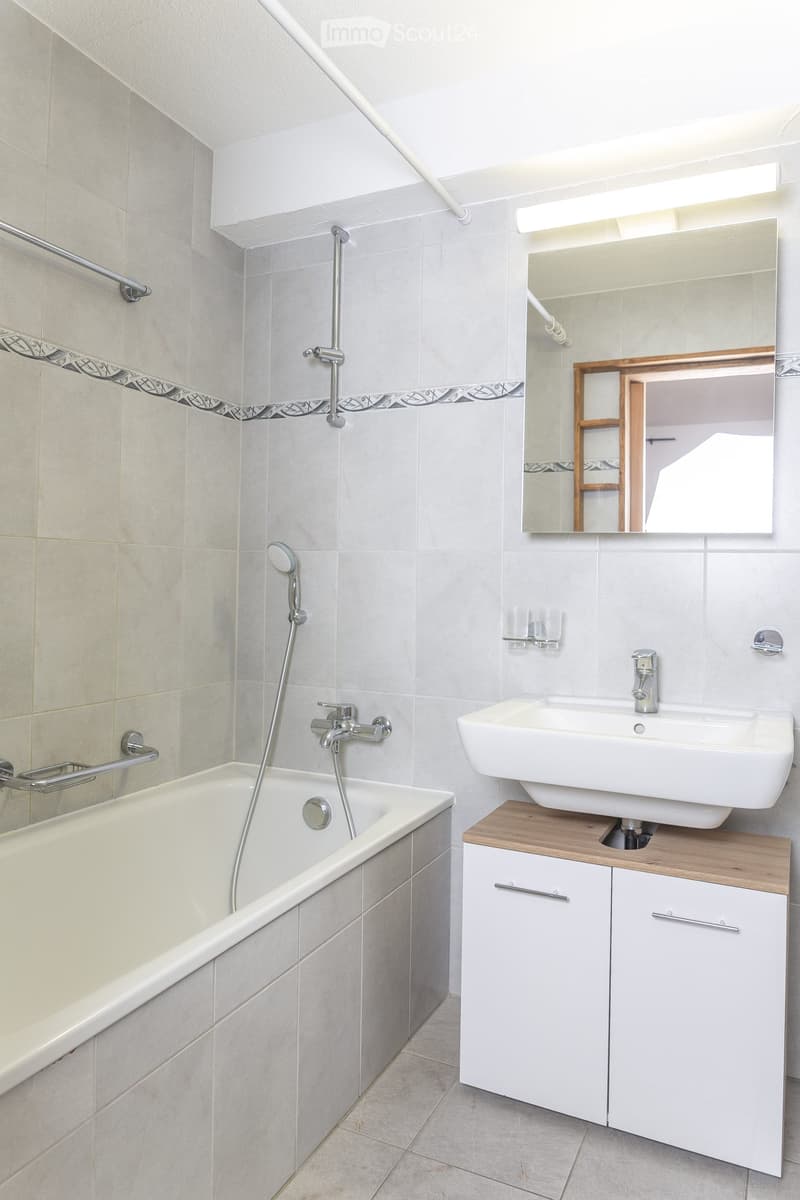 Modern bathroom with a bathtub, sink, mirror, and wooden cabinet. Features a showerhead and faucet above the bathtub.