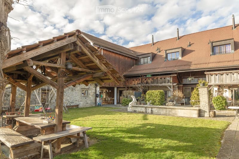A rustic wooden house with a red-tiled roof, surrounded by a well-maintained lawn, wooden benches, and a swing set. The house features a balcony and a stone foundation, with potted plants and outdoor furniture visible near the entrance.