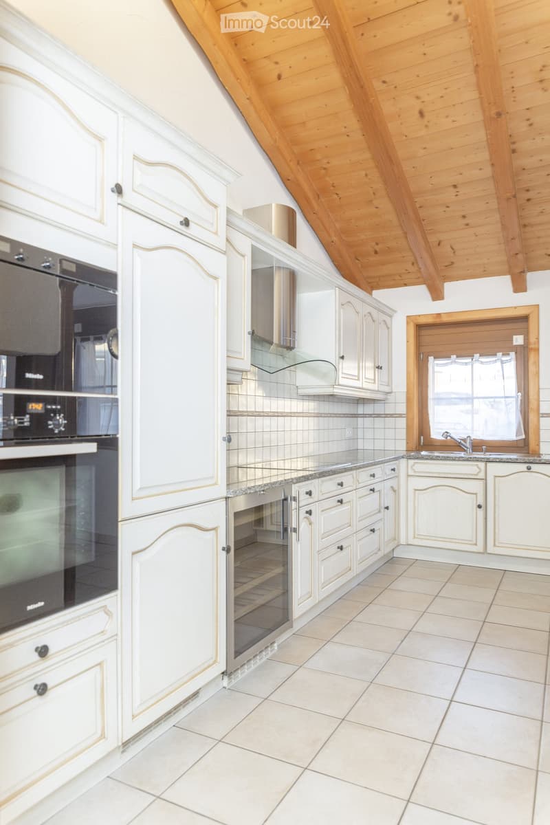 Bright kitchen with white cabinets, a stainless steel oven, a sink with a faucet, and a window with a curtain. The ceiling is wooden with exposed beams.