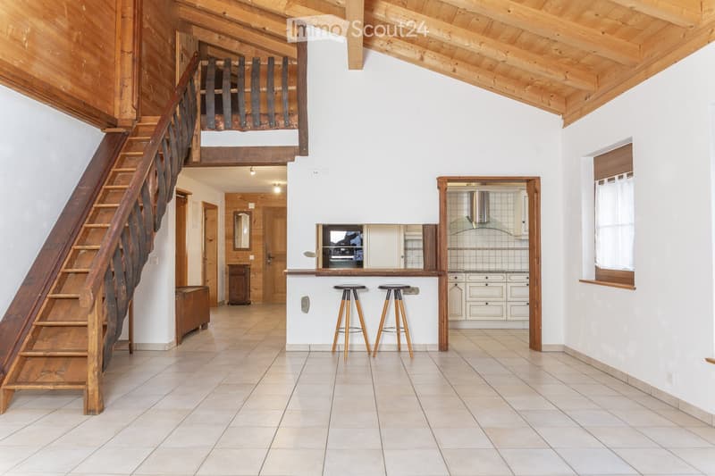 Open-plan living and dining area with a wooden staircase leading to an upper level, a kitchen area with cabinets and a range hood, and two stools at a kitchen island.