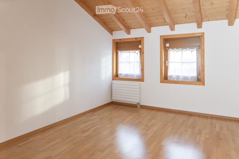 Empty room with wooden flooring, white walls, and two windows with wooden frames and white curtains. A white radiator is mounted on the wall.