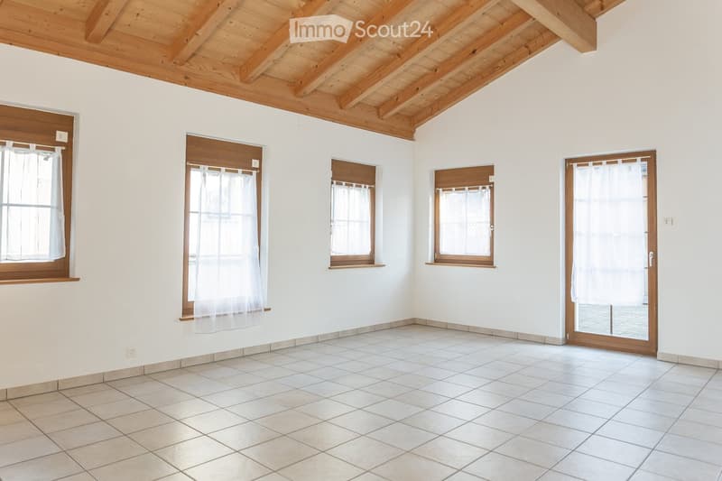 Empty room with white walls, tiled floor, wooden ceiling beams, and multiple windows with sheer curtains. The room features a sliding glass door leading outside.
