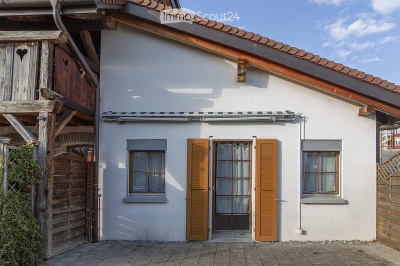 Single-story house with white exterior, wooden shutters, and a tiled roof. Features a covered entrance with a wooden gate and a window with a view of the interior.